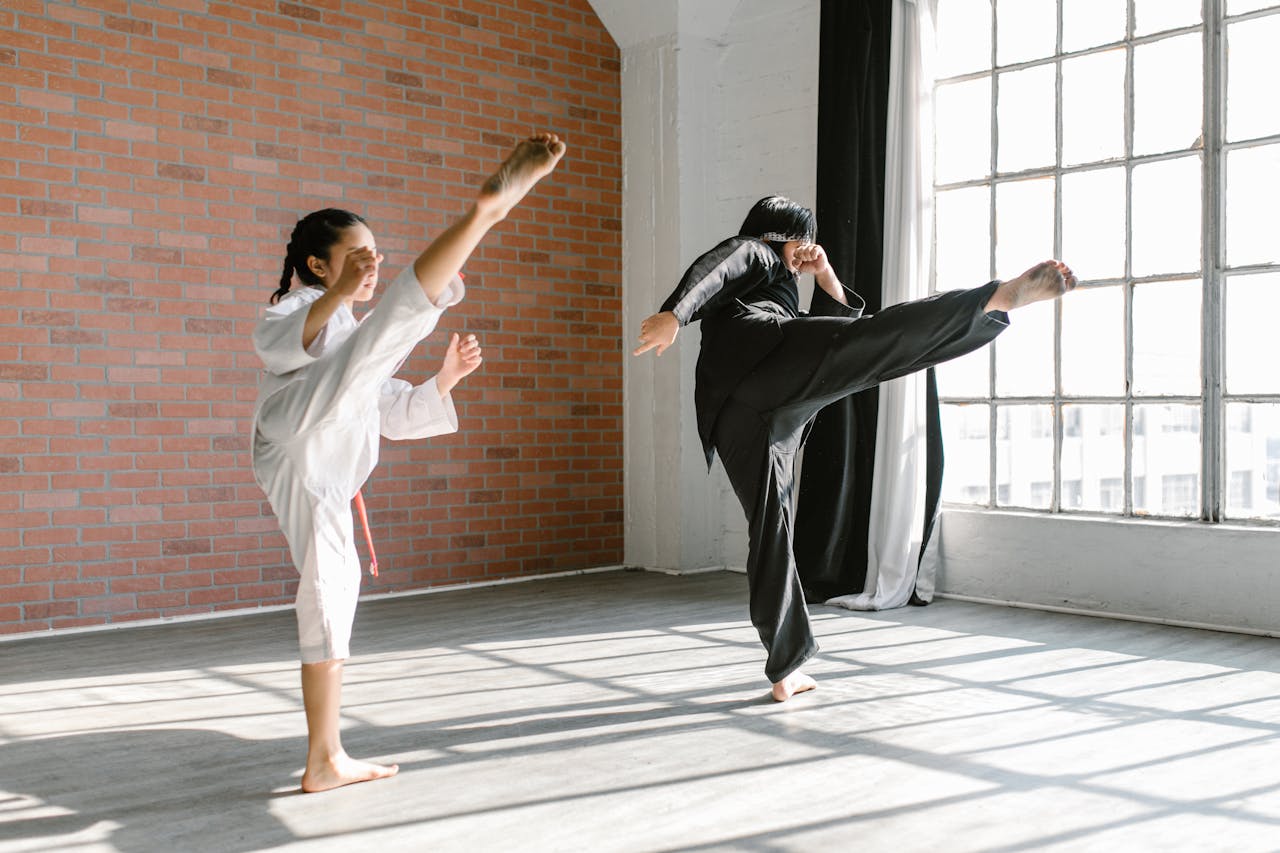 Two martial artists practicing high kicks in a sunlit indoor dojo.